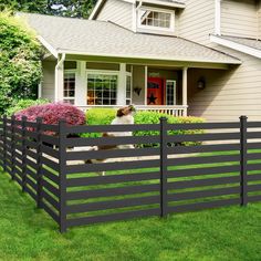 a dog is standing behind a fence in front of a house with grass and bushes