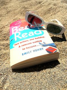 an open book with sunglasses on top of it sitting in the sand next to a pair of sunglasses