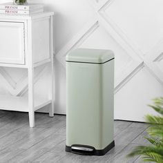 a green trash can sitting next to a white cabinet and plant on top of a wooden floor