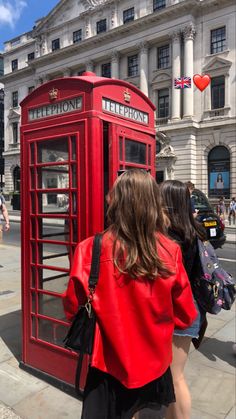 a woman is standing in front of a red phone booth on the sidewalk with her back to the camera