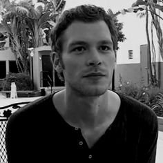 a man standing in front of a fence with palm trees behind him and buildings on the other side