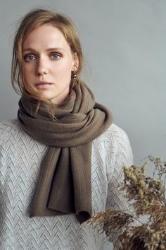 a woman wearing a brown scarf and white sweater is standing in front of a plant