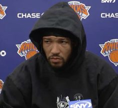 a man in a black hoodie sitting at a table with microphones behind him