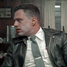 a man in a leather jacket and tie looking off into the distance while sitting down