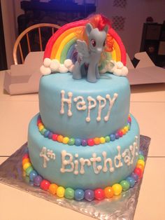 a blue birthday cake with a pony on top and rainbows around the edges that says happy 4th birthday