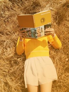 a woman in yellow shirt and white shorts reading a book while standing on dry grass