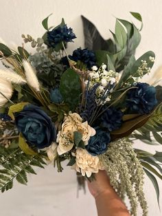 a hand holding a bouquet of flowers and greenery in front of a white wall