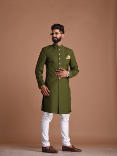 a man in a green sherwa with white pants and brown shoes standing against a brown background