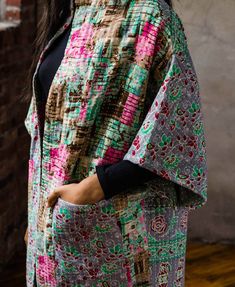 a woman standing in front of a brick wall wearing a multicolored jacket and black top