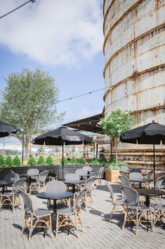 tables and chairs with black umbrellas are set up in front of an old silo