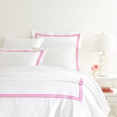 a bed with white sheets and pink trimmings in a bright room, next to a lamp