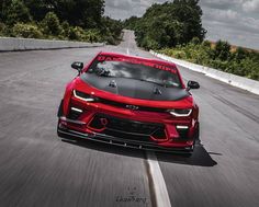 the front end of a red sports car driving on a road with trees in the background