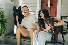 a man and woman are sitting on the porch with their two dogs, one black and one brown