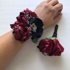 two wrist corsages with red and black flowers on them, one being held by a woman's hand