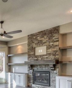 an open concept living room with stone fireplace and built - in shelving units, ceiling fan, and windows