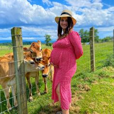 a pregnant woman in a pink dress and hat standing next to a fence with cows