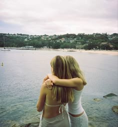 two young women standing on the edge of a body of water, hugging each other