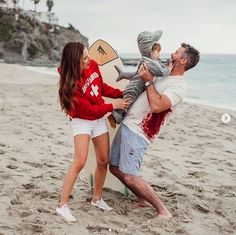 a man and woman holding a baby on the beach with a surfboard in front of them