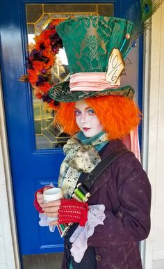 a woman with red hair wearing a green top hat and holding a cup in front of a blue door