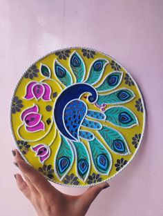 a hand holding a decorative plate with a peacock design on the front and side, against a pink wall