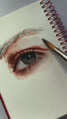 a pencil drawing of an eye with red and black liner on paper next to a spiral notebook