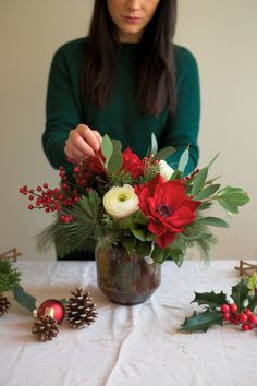 a woman arranging flowers in a vase on top of a table with pine cones and holly