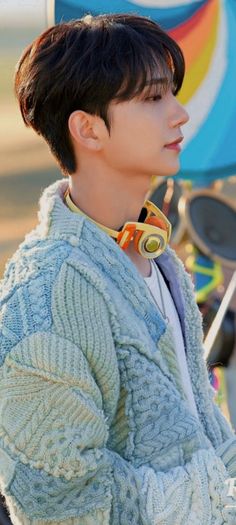 a young man with short hair wearing a sweater and scarf, standing in front of a colorful kite