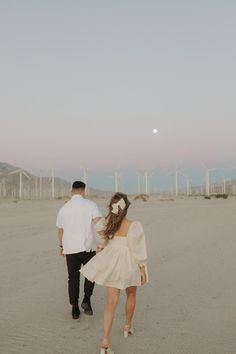 a man and woman walking in the sand with wind mills behind them at sunset or dawn