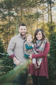 a man and woman holding a baby standing in front of christmas trees with their arms around each other