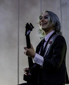 a woman with grey hair holding a guitar in her right hand and smiling at the camera