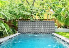 an outdoor swimming pool surrounded by lush green trees and grass, with a tiled wall in the background