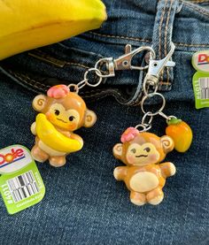 two monkey keychains sitting on top of a pair of jeans with bananas and an apple