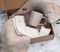 an open box with a blanket, mug and spoon in it on a table next to flowers