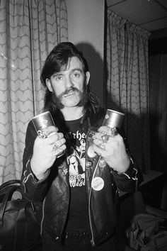 a man with long hair holding two cups in his hands and looking at the camera