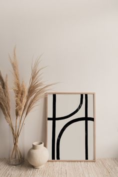 a white vase sitting next to a painting on top of a wooden table near a plant