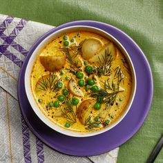 a bowl of soup with potatoes and peas on a purple plate next to a spoon