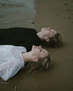 two people laying on the ground in the sand at the beach with their heads close to each other