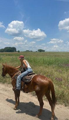 a woman riding on the back of a brown horse down a dirt road next to a lush green field