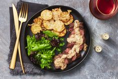 a black plate topped with meat and veggies next to a glass of wine