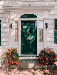 a green front door on a white house with red flowers in the foreground and black shutters