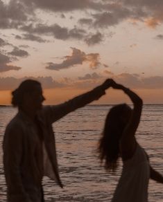 a man and woman standing on top of a beach next to the ocean at sunset