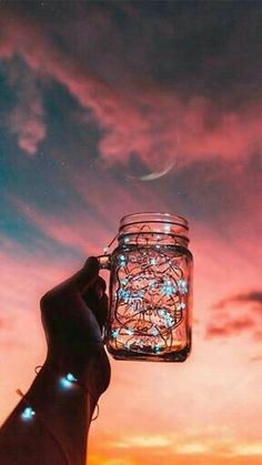 a person holding a mason jar with string lights in it at night under a colorful sky