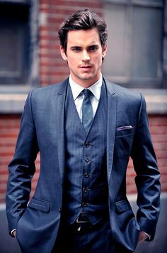 a man wearing a suit and tie standing in front of a brick building with his hands in his pockets