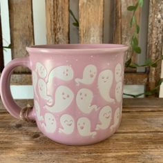 a pink mug with ghost faces on it sitting on a wooden table next to a plant