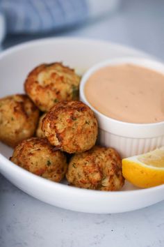 a white bowl filled with crab cakes next to a lemon wedge and dipping sauce on the side