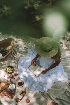 a woman in a white dress and hat sitting on a blanket with an open book