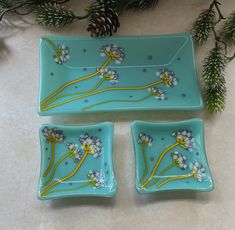 three blue dishes with flowers painted on them and pine branches in the background, sitting on a table