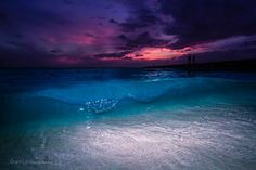 an ocean wave is coming in to the shore at night with purple and blue colors