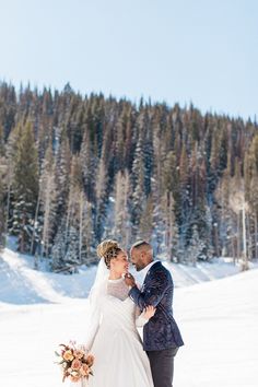 a bride and groom standing in the snow with trees in the background