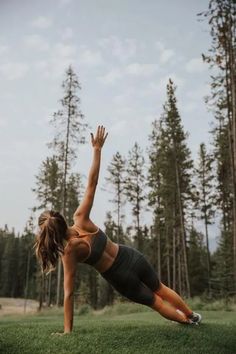 a woman is doing yoga on the grass in front of some trees and pine trees